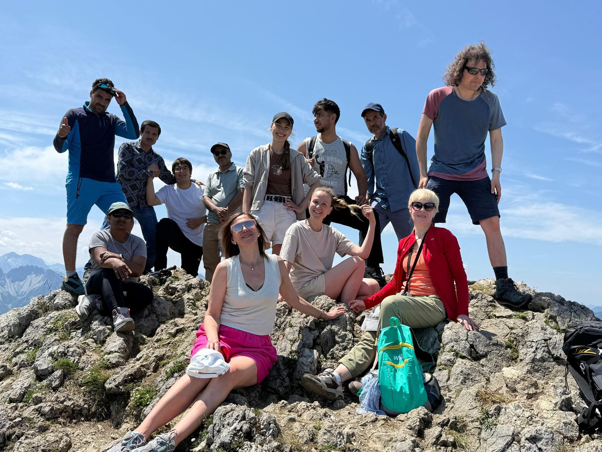 Gruppe von elf Personen auf felsigem Gipfel, einige sitzen, andere stehen, Berglandschaft im Hintergrund