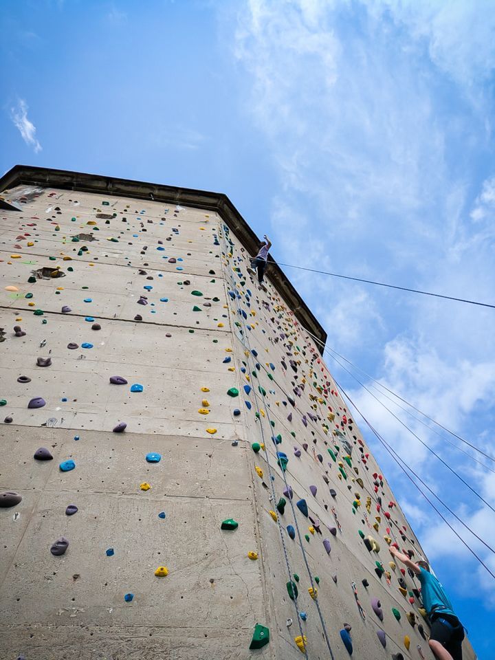 Es ist die Außenkletteranlage zu sehen. Eine Person wird gerade abgelassen und hebt einen Arm.