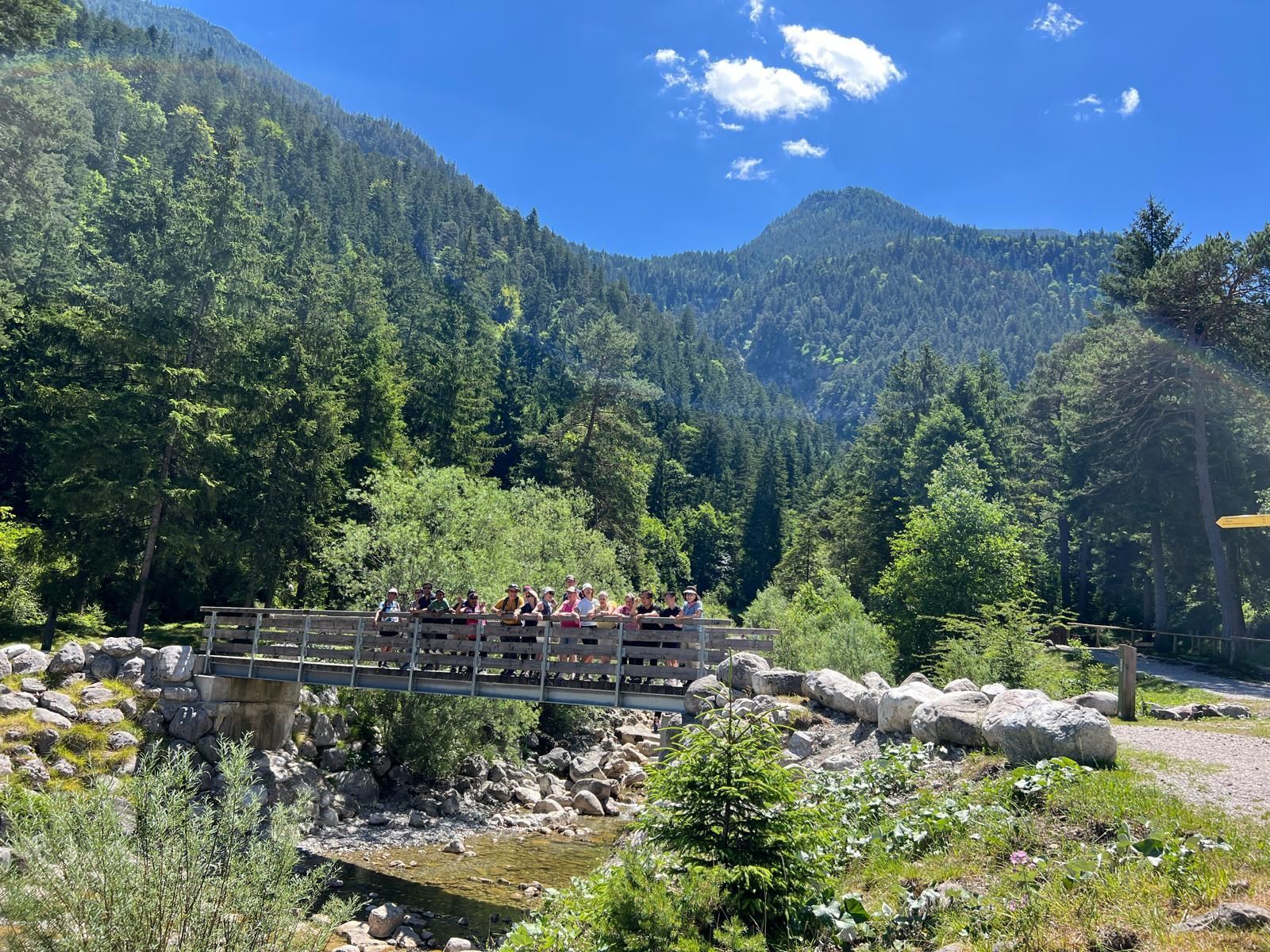 Eine Gruppe von Menschen steht auf einer Holzbrücke über einem kleinen Fluss, umgeben von grünen Bäumen und Bergen. Der Himmel ist blau mit einigen Wolken.