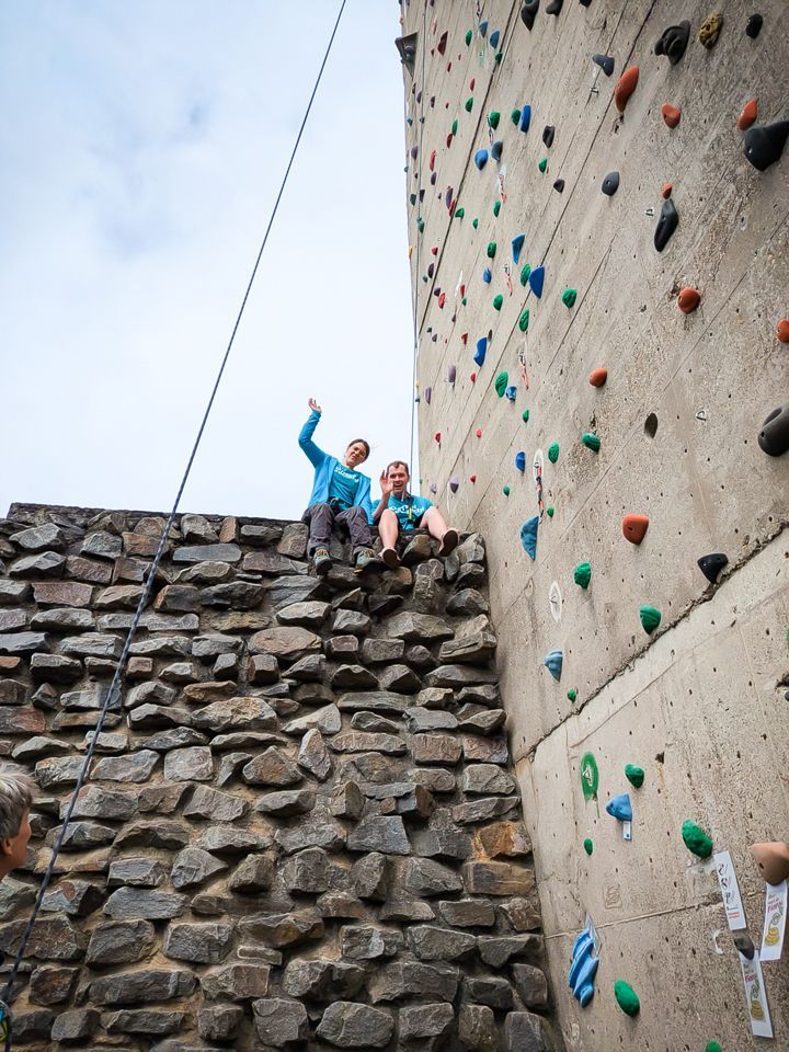 Es sind zwei Personen auf einer Steinmauer neben der Außenkletteranlage zu sehen. Sie winken in die Kamera.