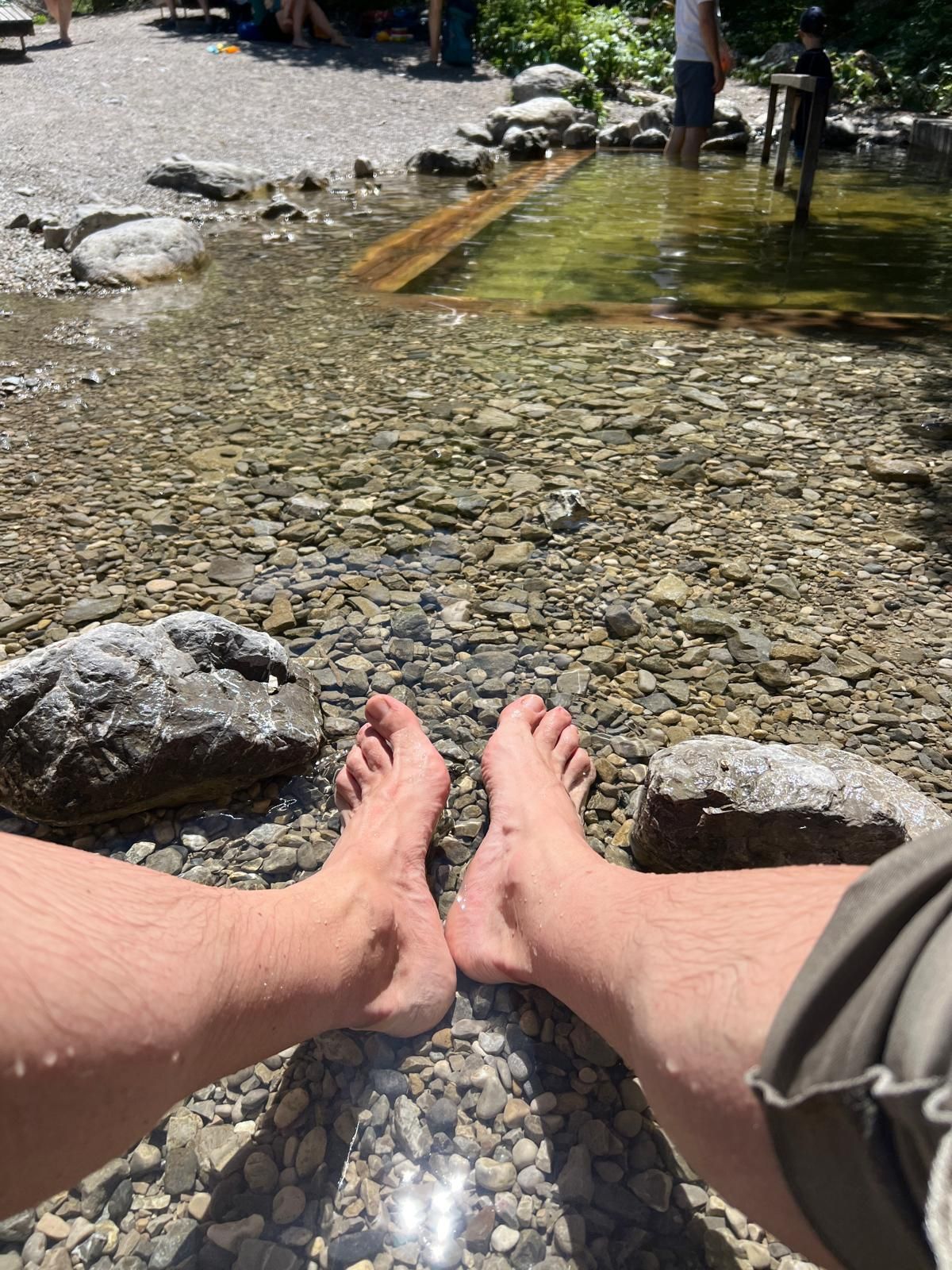 Zwei nackte Füße stehen im flachen Wasser eines Baches, umgeben von kleinen Steinen und Felsen. Im Hintergrund sind verschwommene Personen und Bäume sichtbar.