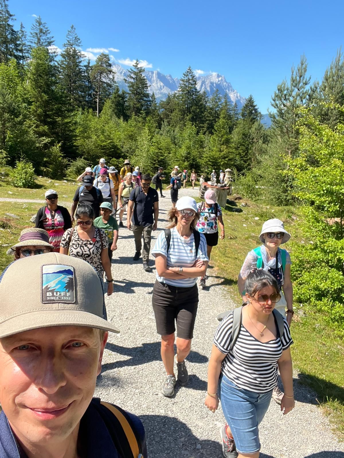 Gruppe von Wanderern auf einem Waldweg neben einem Feld mit Bergen im Hintergrund unter bewölktem Himmel