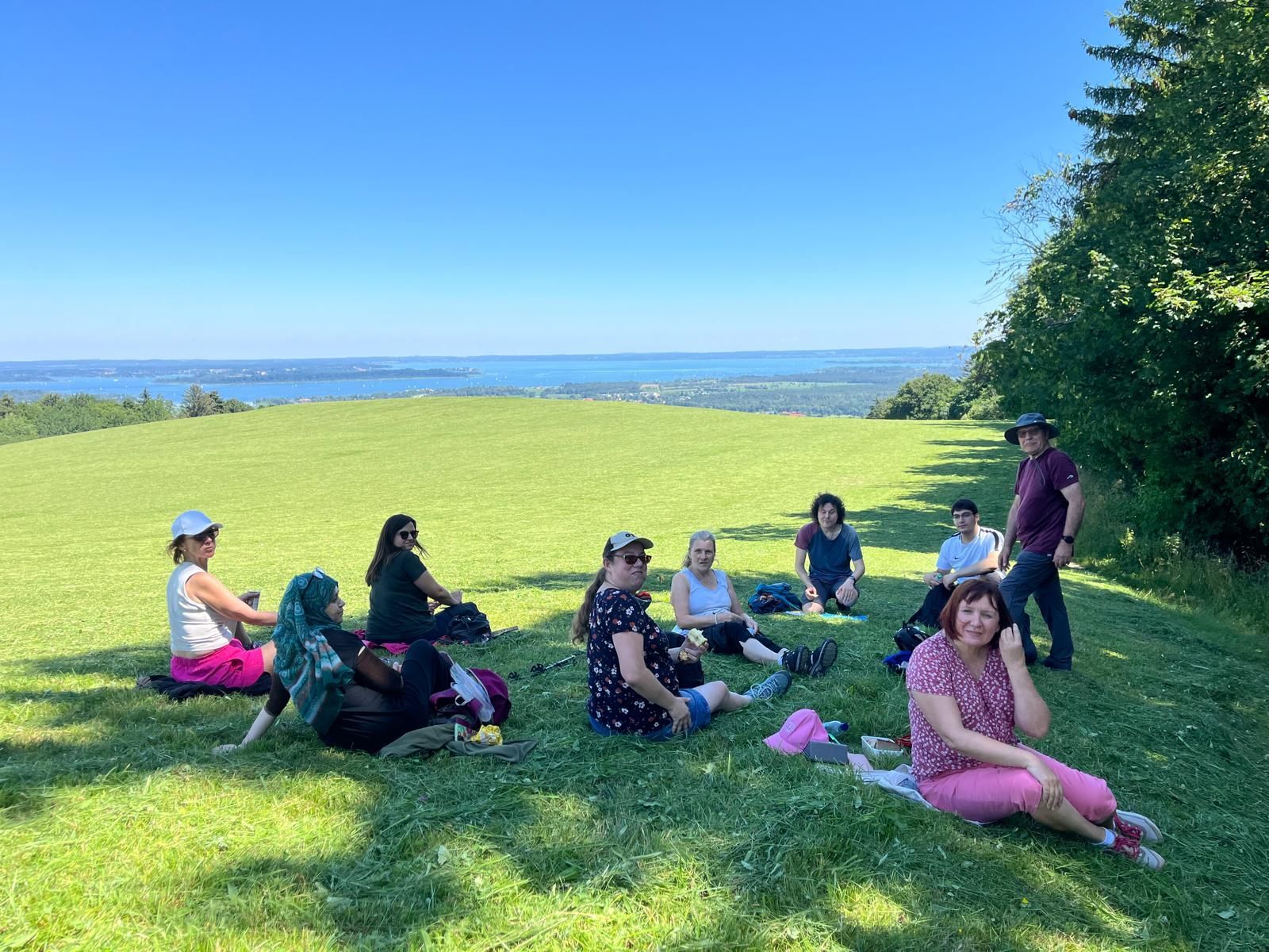 Eine Gruppe von acht Personen sitzt auf einem Hügel in einer grünen Wiese mit Blick auf einen See. Der Himmel ist klar und blau.