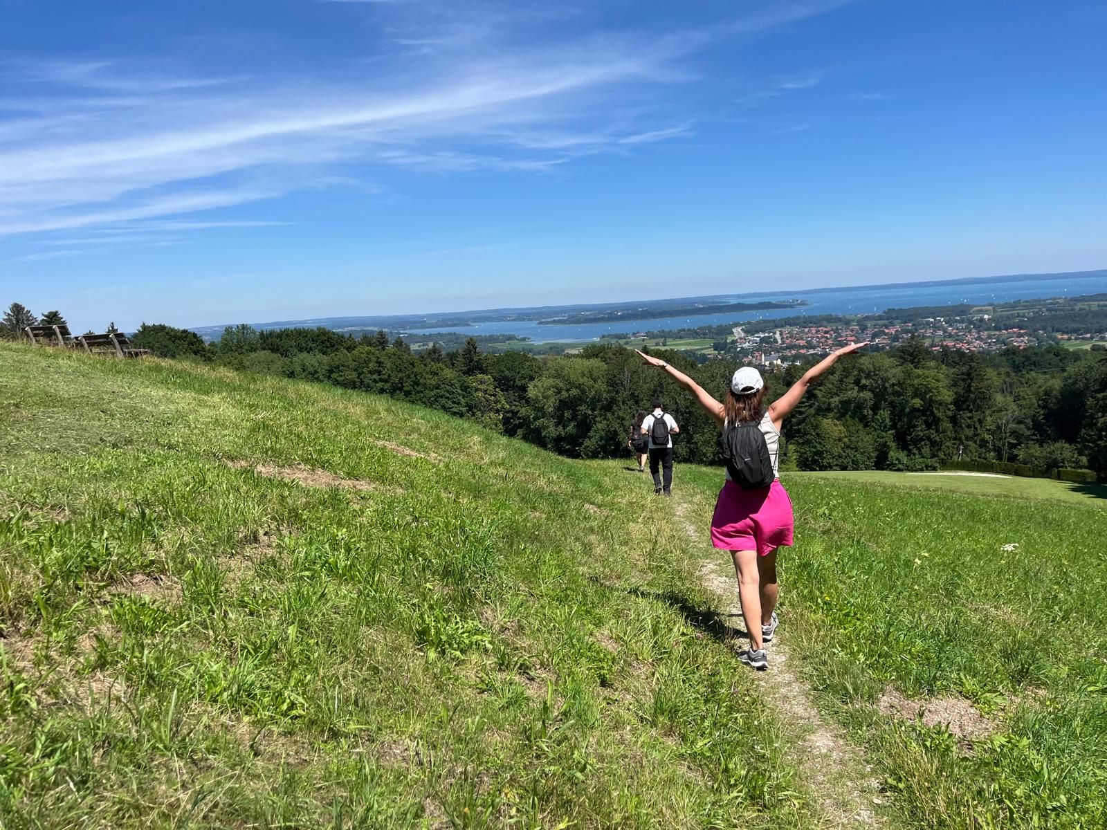 Frau mit weißer Kappe und pinkem Rock wandert auf einem Pfad durch eine grüne Wiese mit Blick auf einen See und bewaldete Hügel