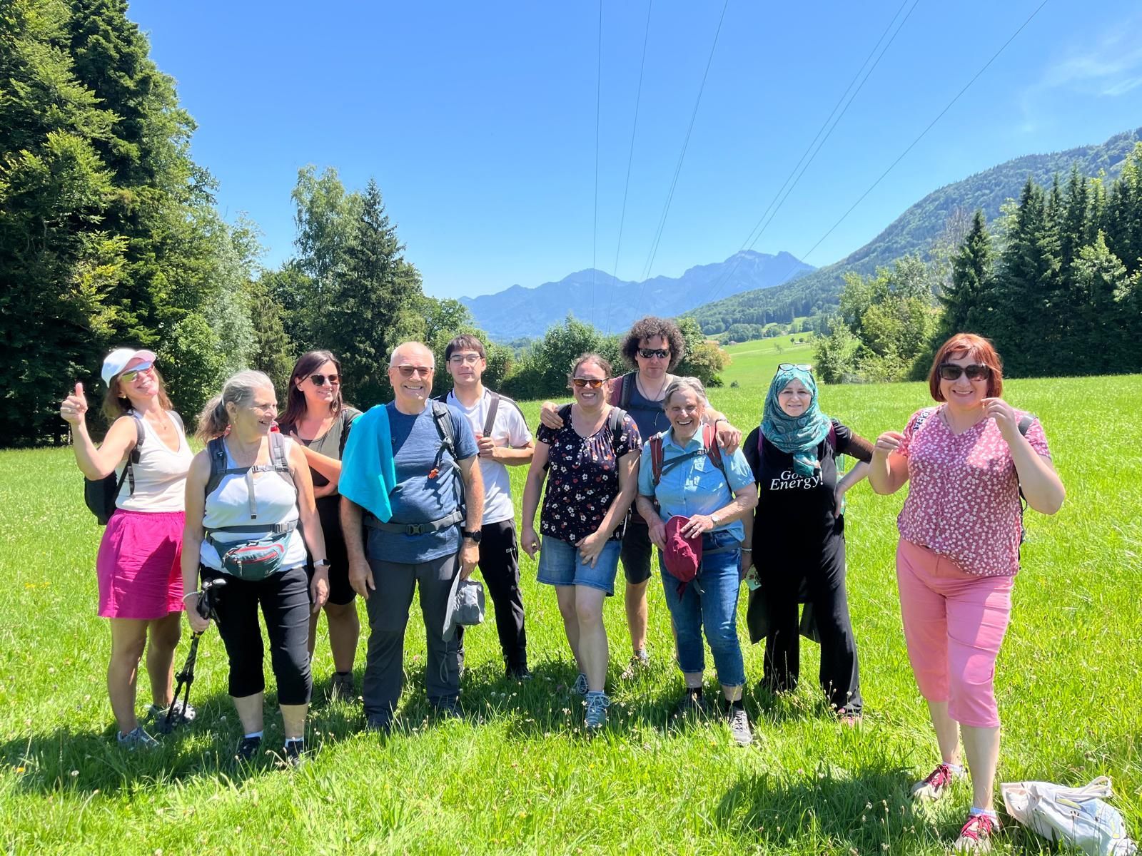 Gruppe von zehn Personen steht auf einer grünen Wiese vor Bergen unter blauem Himmel, einige tragen Wanderkleidung und Rucksäcke.