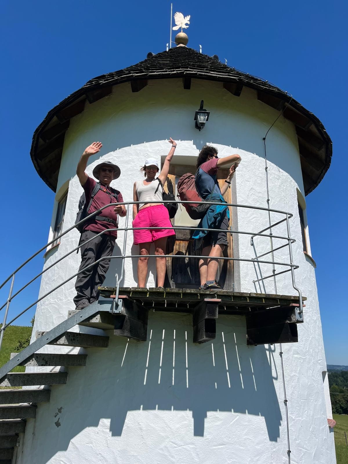 Drei Personen stehen auf dem Balkon eines runden weißen Turms mit schwarzem Dach, zwei winken in die Kamera.