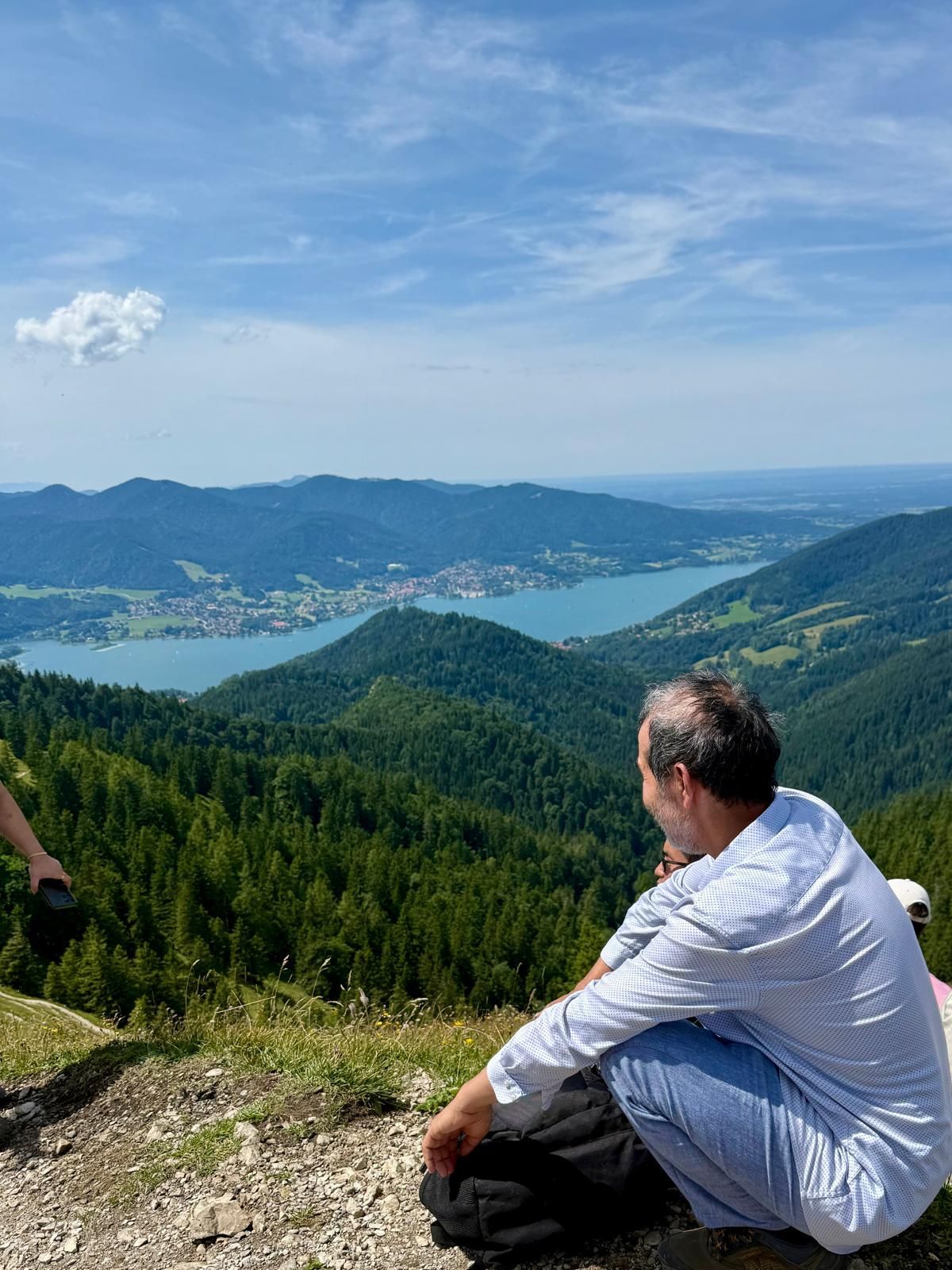 Mann in weißem Hemd sitzt auf einem Berg mit Blick auf bewaldete Hügel und einen See in der Ferne