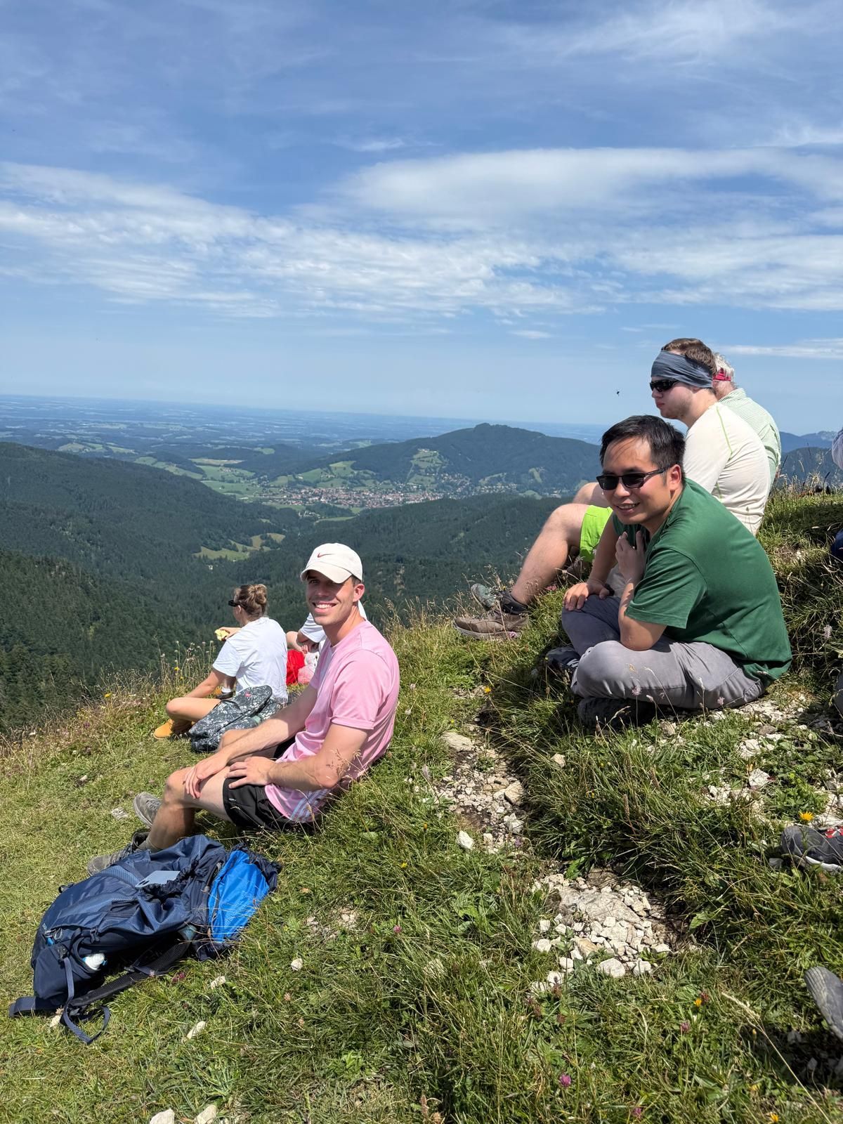 Fünf Personen sitzen auf einer Bergwiese und blicken auf ein Tal mit Dörfern und bewaldeten Hügeln unter blauem Himmel