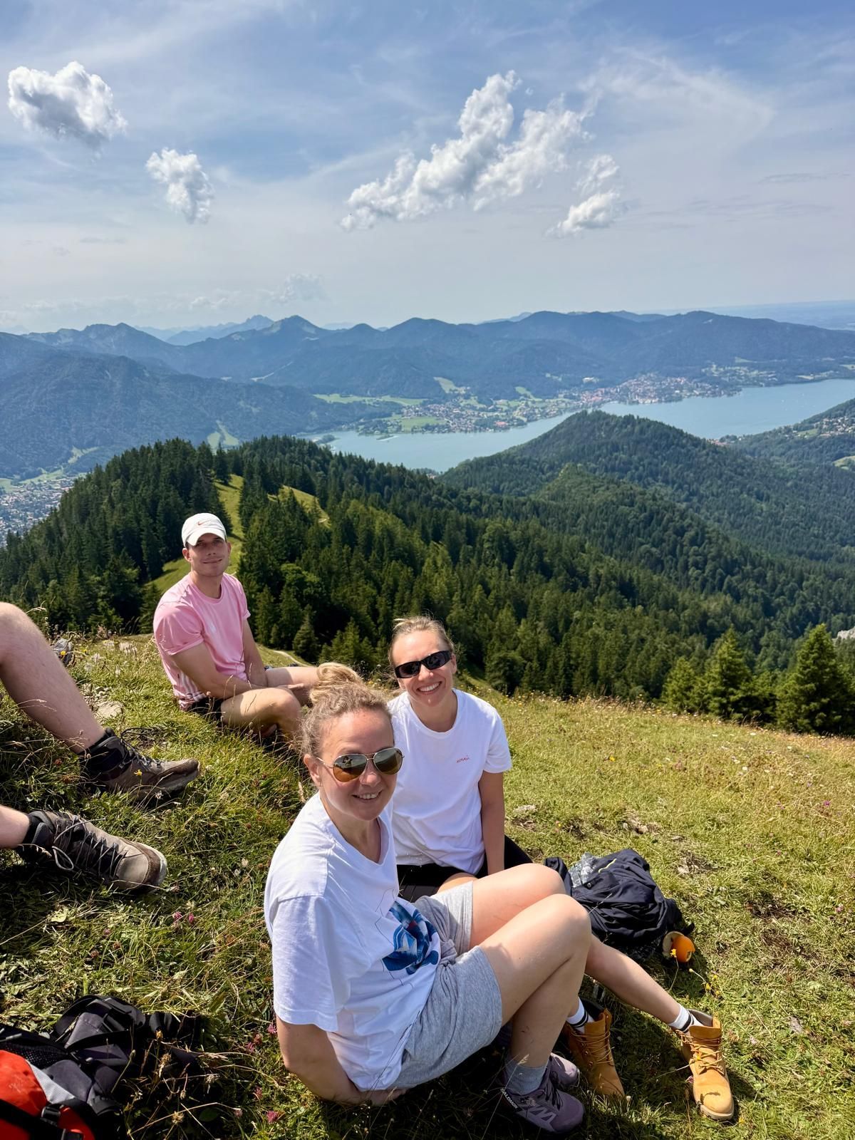 Drei Personen sitzen auf einer Bergwiese mit Blick auf bewaldete Hügel und einen großen See unter blauem Himmel mit Wolken