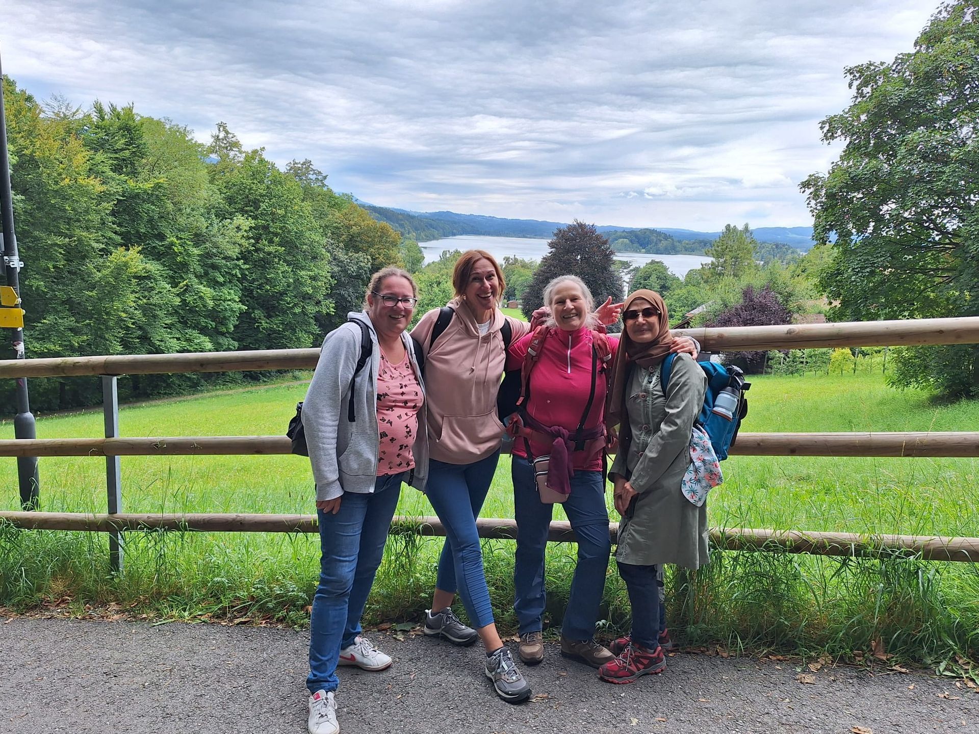Vier Personen stehen auf einem Weg vor einer grünen Wiese mit Bäumen im Hintergrund. Zwei Frauen tragen Rucksäcke, eine hat eine graue Jacke und die andere eine rote. Die Umgebung ist bewaldet und hügelig.