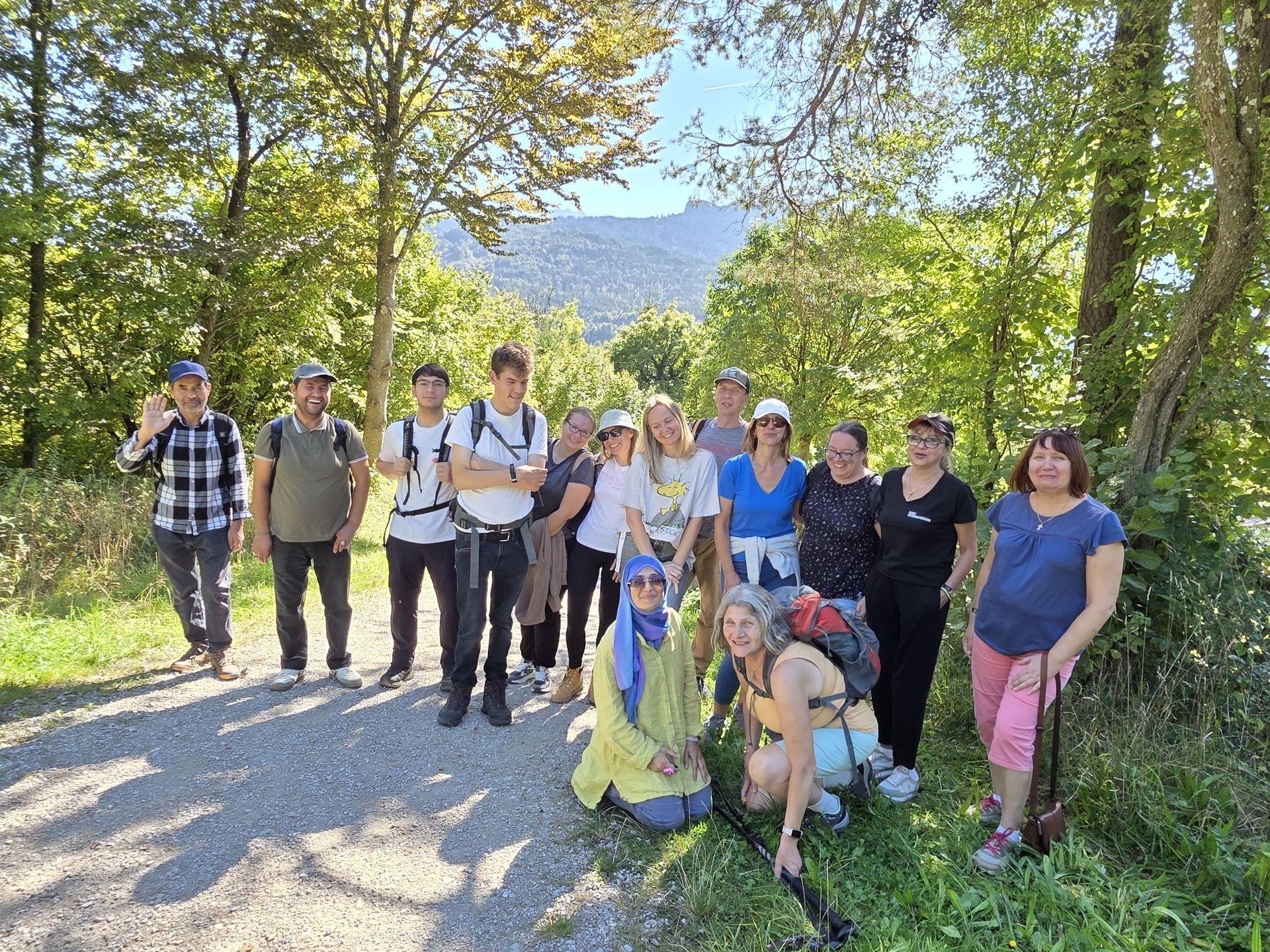 Gruppe von Menschen in Outdoor-Kleidung auf einem Waldweg, umgeben von Bäumen und Bergen im Hintergrund.