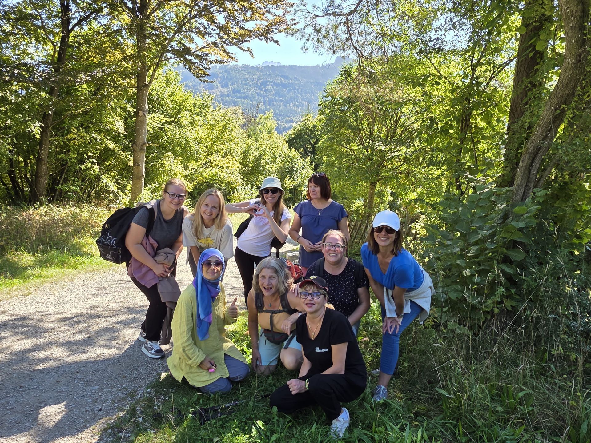 Gruppe von Menschen in Outdoor-Kleidung posiert auf einem Waldweg, umgeben von Bäumen und Bergen im Hintergrund.