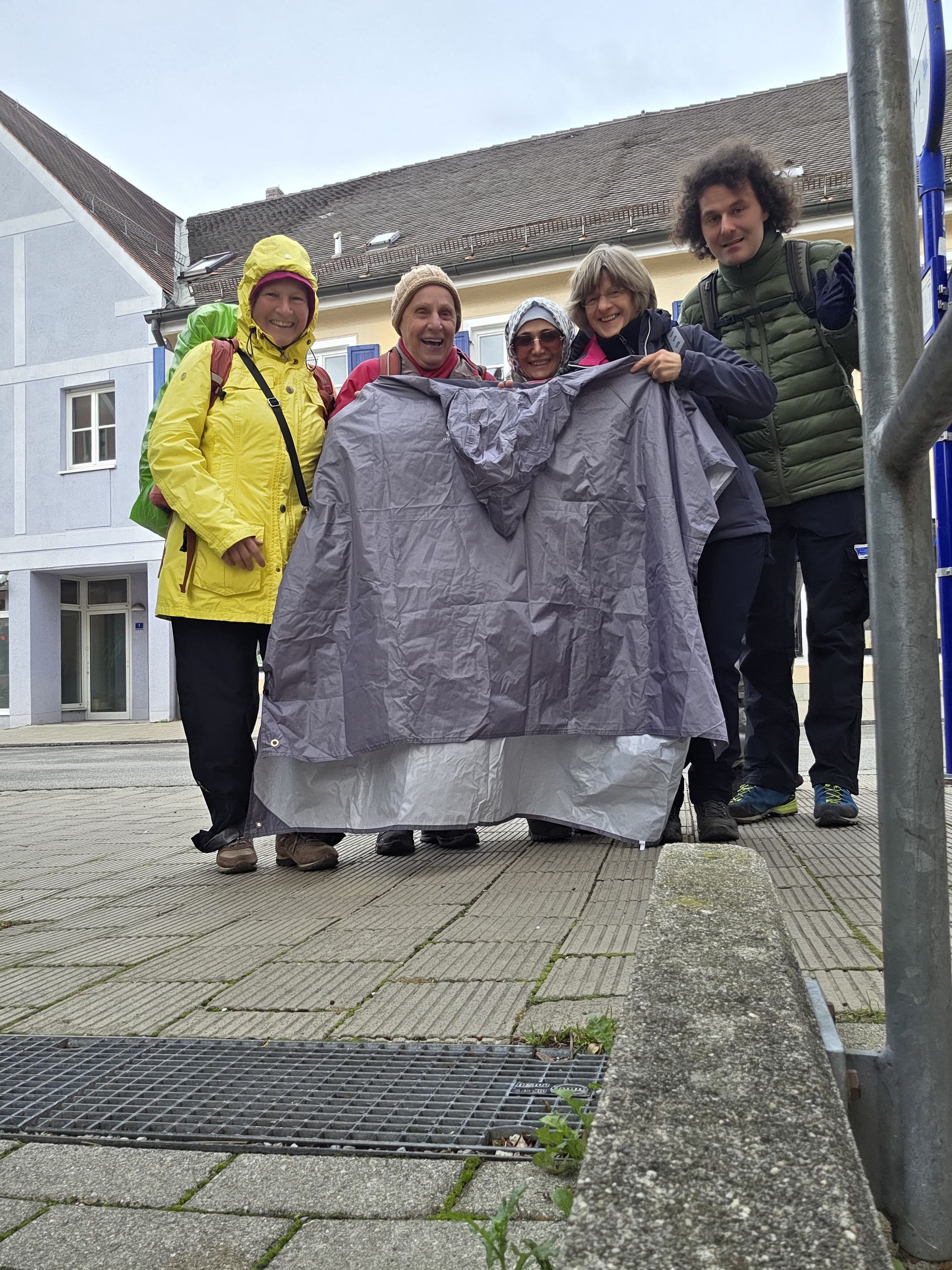 Fünf Personen stehen auf einem gepflasterten Platz, eine trägt eine gelbe Regenjacke, drei halten einen Regenumhang.