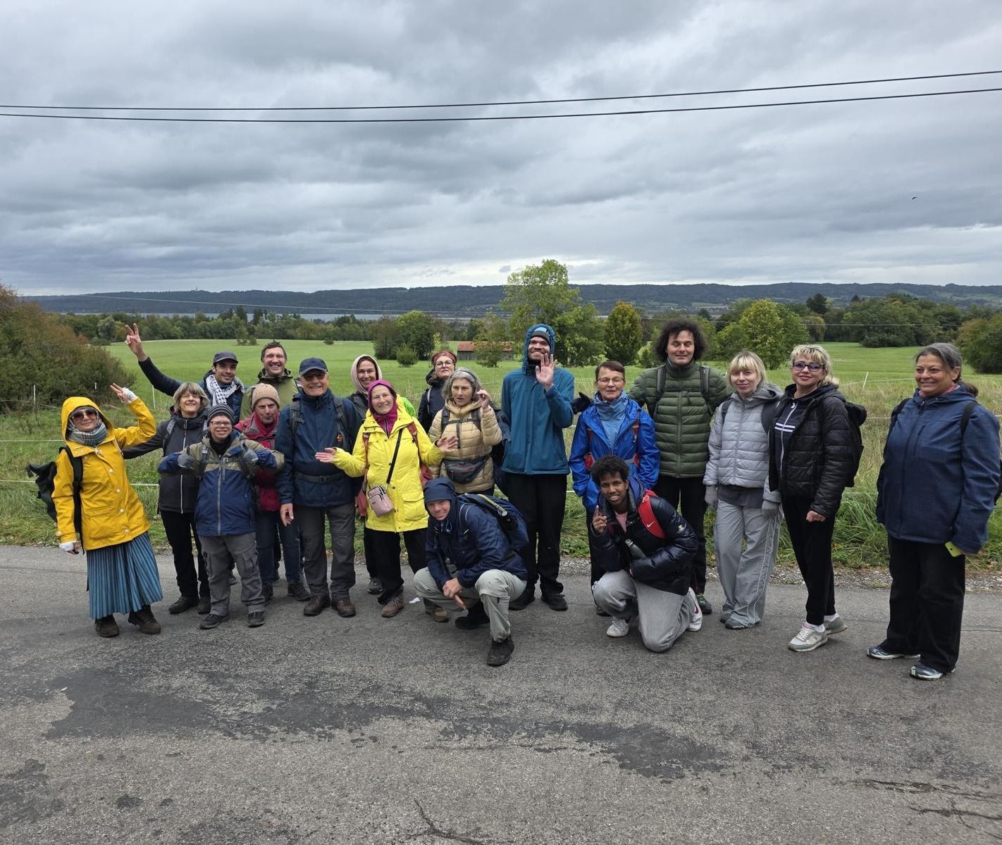 Gruppe von Menschen in Outdoor-Kleidung steht auf einer Straße vor einer Landschaft mit Bäumen und Hügeln unter bewölktem Himmel.
