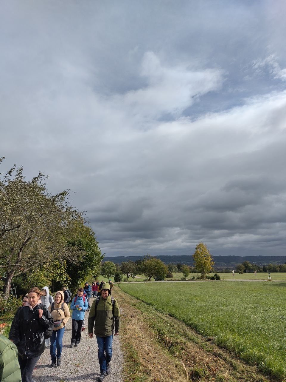 Wandergruppe auf einem schmalen Weg neben einer Wiese unter bewölktem Himmel mit Bäumen am Wegesrand.