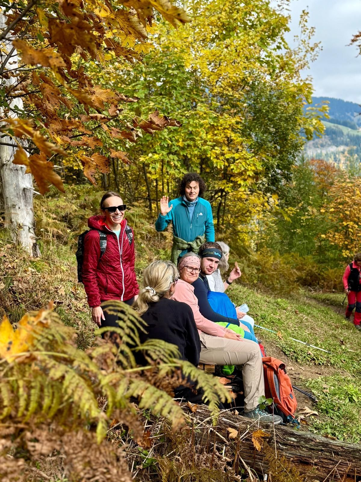 Gruppe von Menschen in herbstlicher Waldlandschaft, einige sitzen auf Baumstamm, andere stehen, umgeben von gelb-grünem Laub.