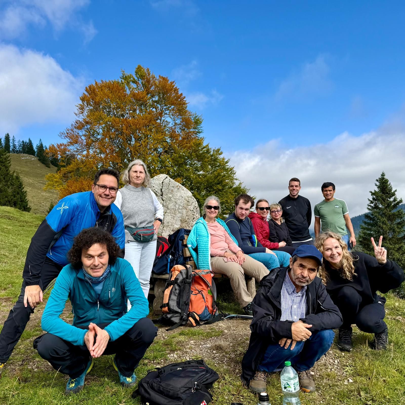 Gruppe von Wanderern in Outdoor-Kleidung sitzt und steht auf einer Wiese mit Rucksack und Wasserflasche, im Hintergrund ein Baum mit herbstlichen Blättern und ein
bewaldeter Hügel.