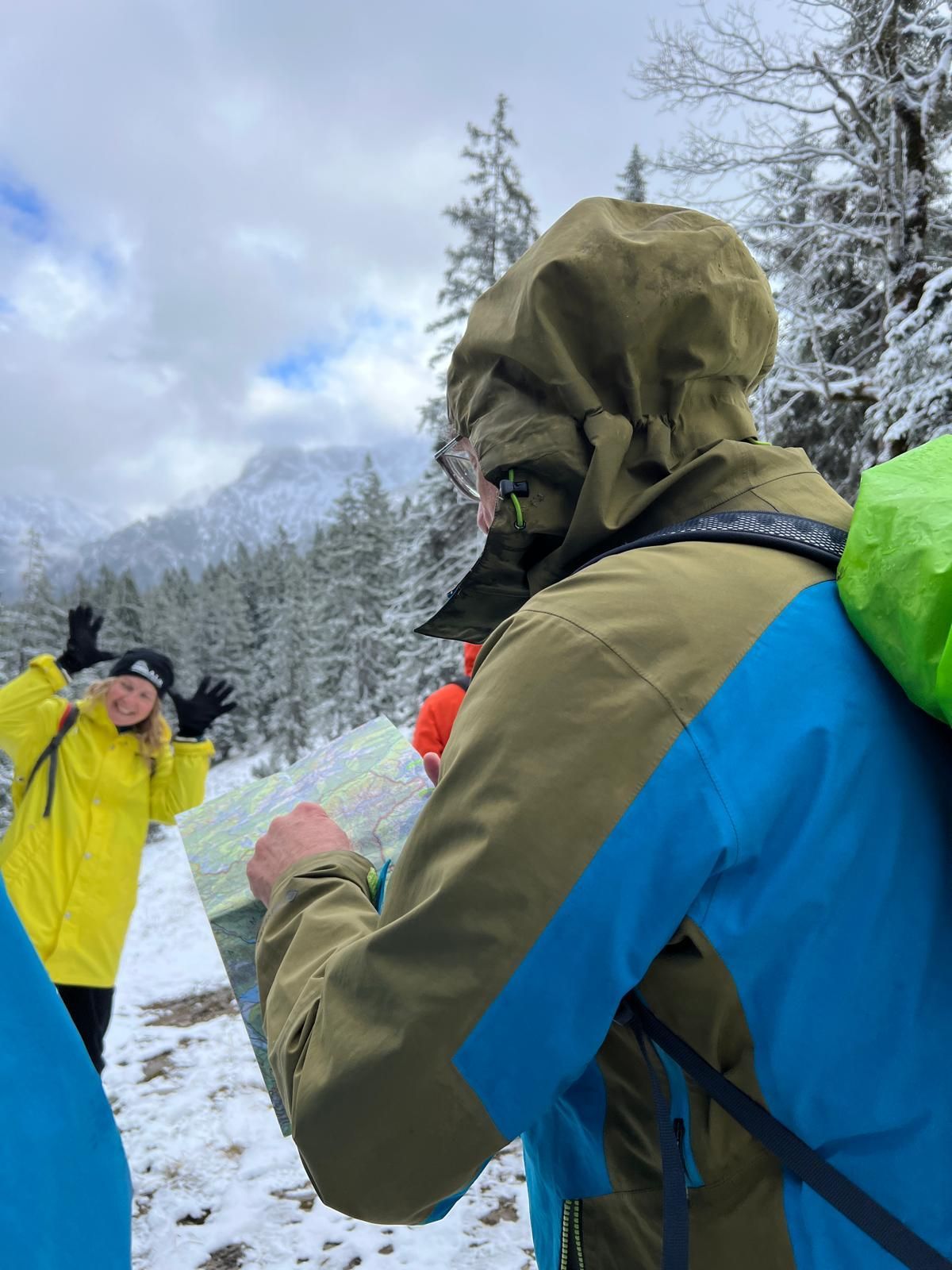 Person in blau-grüner Outdoorjacke mit Kapuze hält eine Karte, im Hintergrund verschneite Bäume und eine weitere Person in gelber Jacke mit erhobenen Händen.