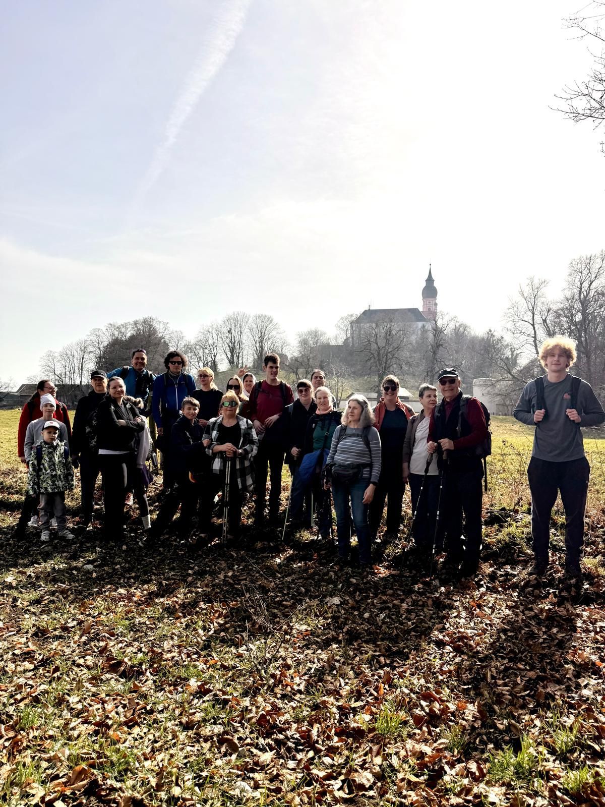 Gruppe von Menschen unterschiedlichen Alters steht auf einem mit Laub bedeckten Feld vor einer Kirche auf einem Hügel im Hintergrund