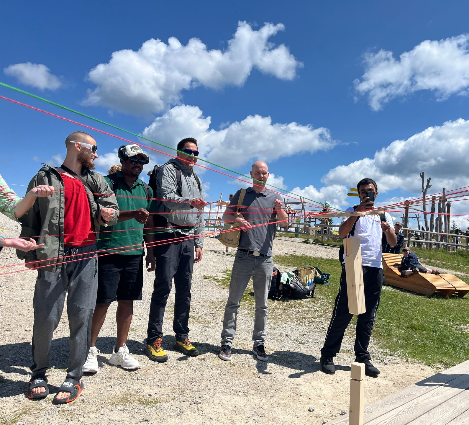 Gruppe von fünf Personen steht auf Holzplanken im Freien und hält nur mit farbigen Schnüre ein Holztablett. An dem Tablett hängt unten ein Haken, in den ein Holzpflock eingehängt ist. Die Gruppe versucht, nur mit Hilfe der Schnüre den eingehängten Pflock auf einen anderen Pflock zu stellen, der senkrecht auf dem Boden steht.