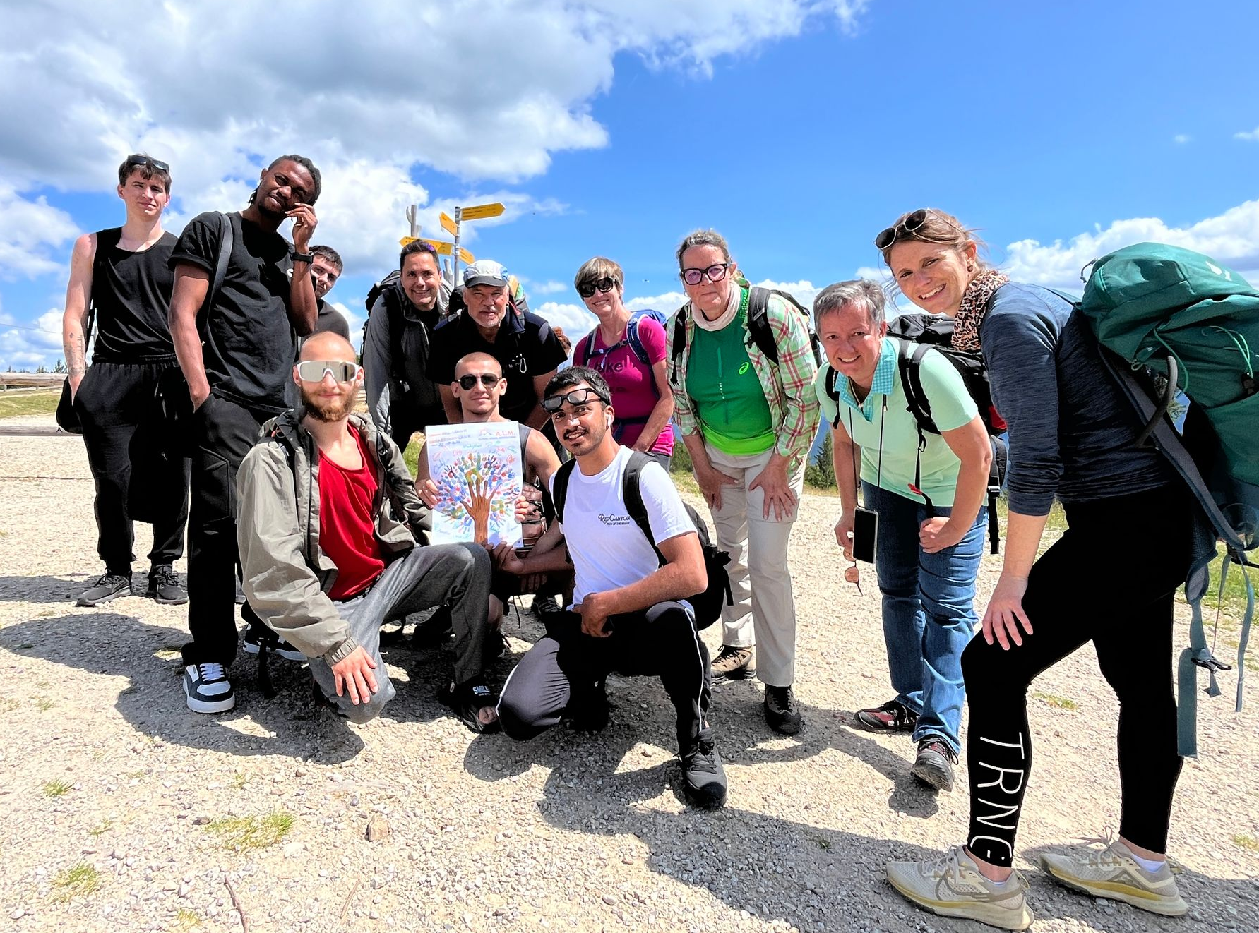 Gruppenfoto von 24 Personen, darunter Erwachsene und Kinder, in einer Berglandschaft mit schneebedeckten Gipfeln im Hintergrund. Ein Hund steht neben einem Kinderwagen.