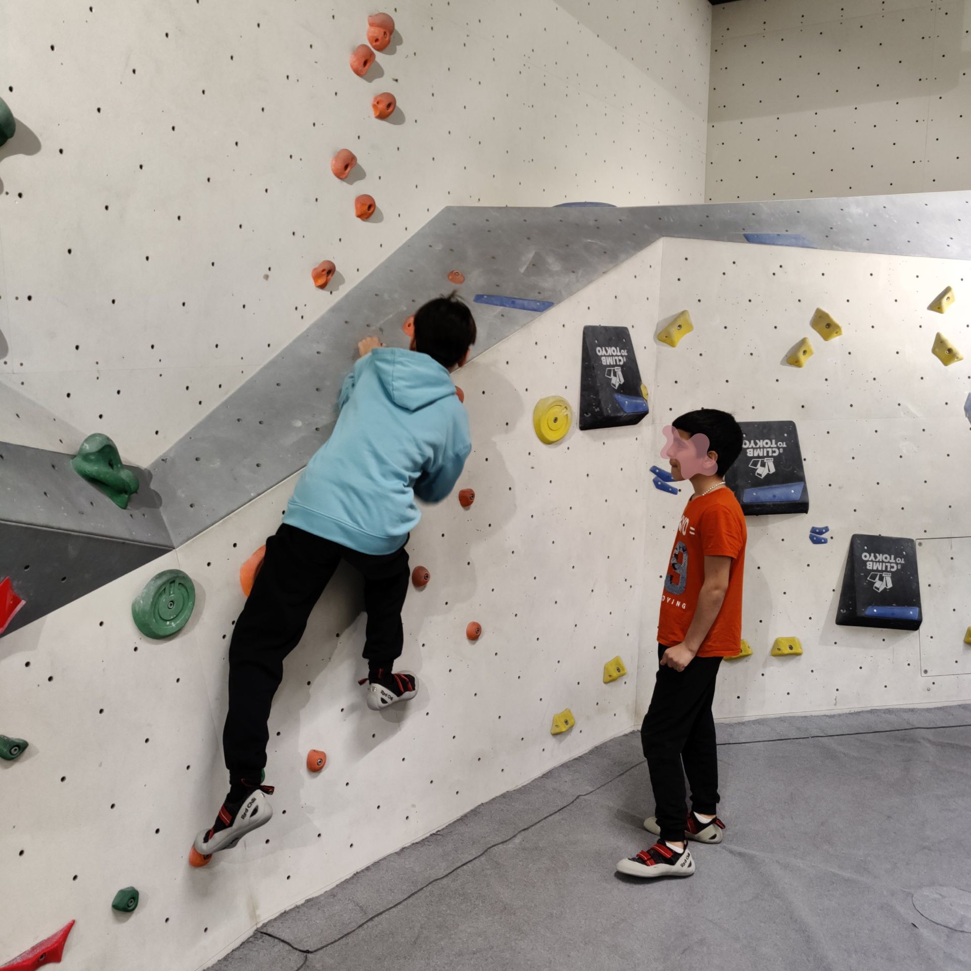 Kind in hellblauer Kapuzenjacke klettert an einer Kletterwand mit bunten Griffen, daneben steht ein weiteres Kind in orangefarbenem T-Shirt