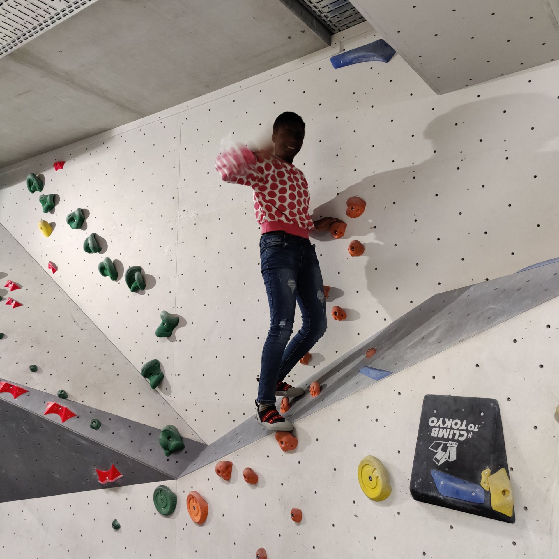 Junge Person klettert an einer Indoor-Kletterwand mit bunten Griffen, trägt rote Pullover mit weißen Punkten und blaue Jeans