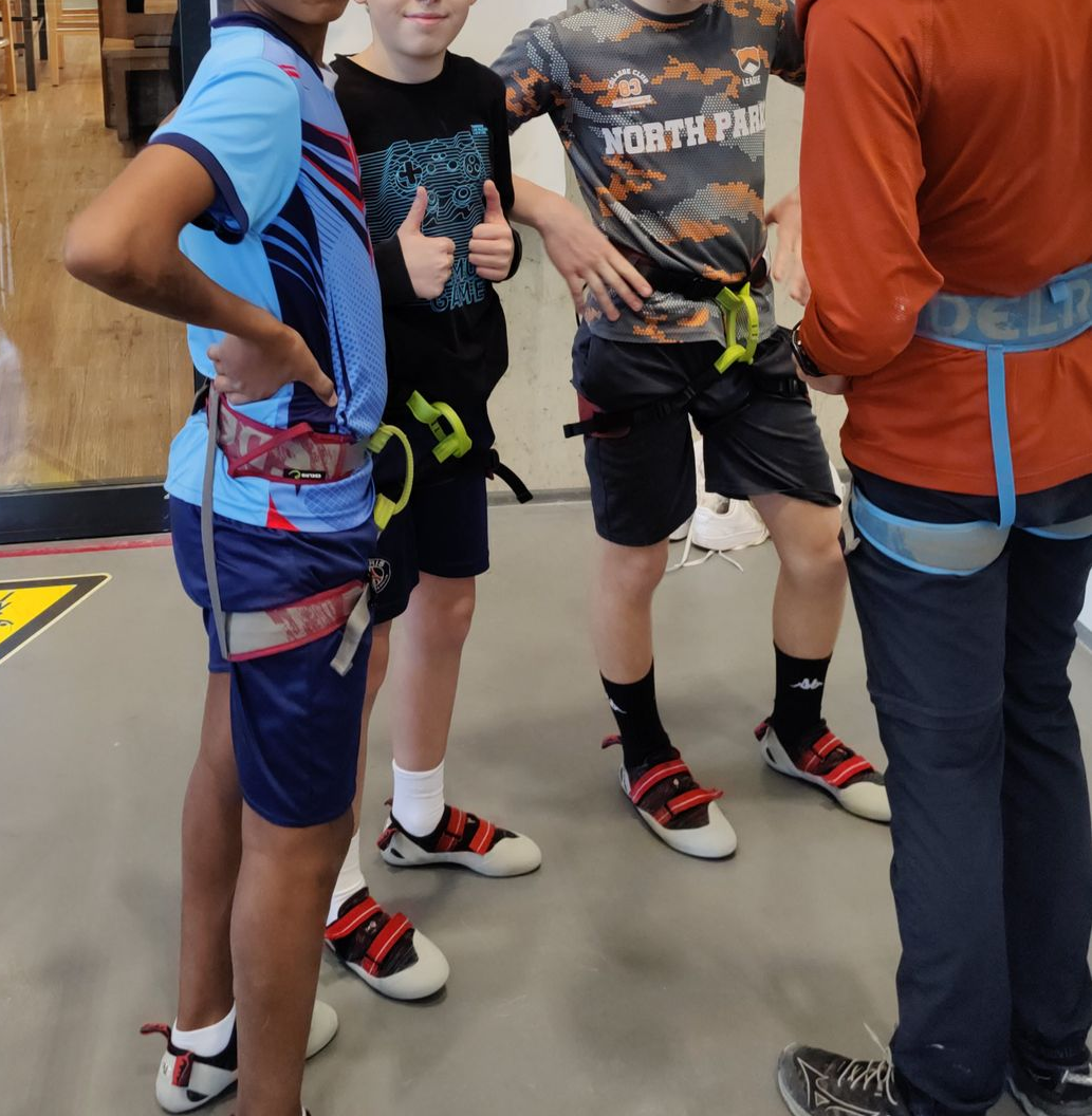 Drei Jungen in Sportkleidung und Kletterschuhen stehen in einer Halle vor einer Wand mit Klettergriffen, eine Person in orangefarbenem Oberteil steht daneben