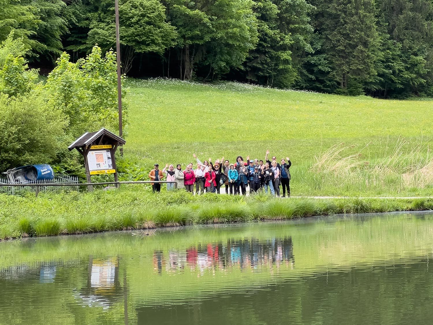 Eine Gruppe von etwa 25 Personen steht am Ufer eines kleinen Sees und winkt. Im Hintergrund sind Bäume und eine Wiese sichtbar. Die Gruppe trägt bunte Kleidung.