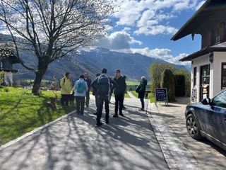 Gruppe von sechs Personen steht auf einem Weg neben einem Haus mit Bergpanorama und blauem Himmel im Hintergrund
