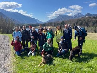 Gruppe von Wanderern mit Rucksäcken und Wanderstöcken auf einer Wiese vor Bergen und blauem Himmel