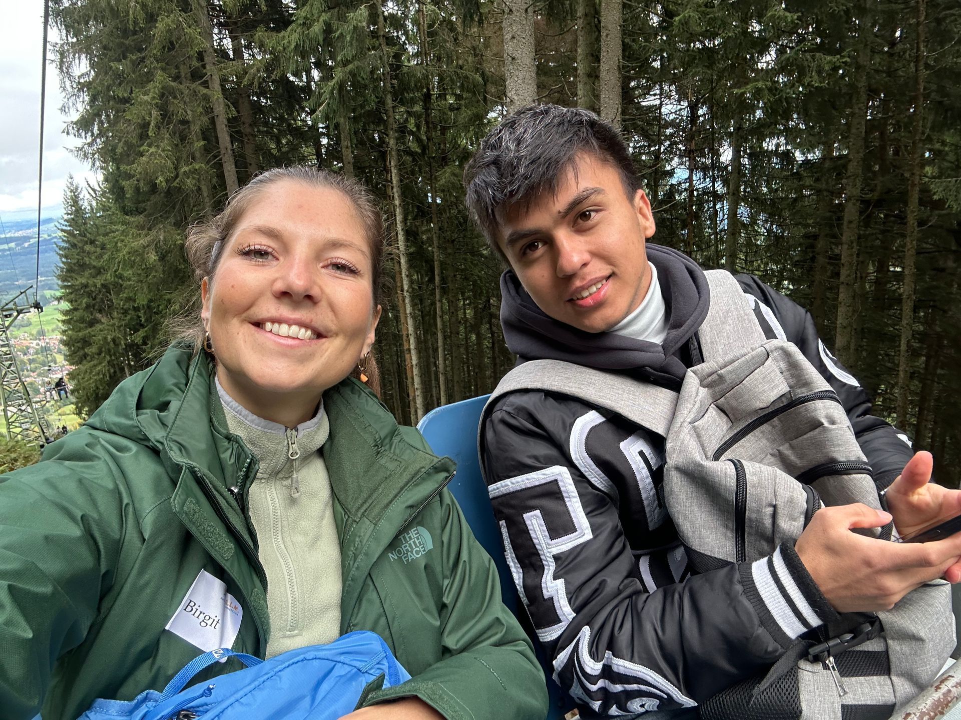 Das Bild zeigt eine Frau und einen Mann, die nebeneinander in einem Sessellift sitzen. Im Hintergrund sind Bäume und eine Landschaft zu sehen. Beide lächeln und scheinen die Fahrt zu genießen.