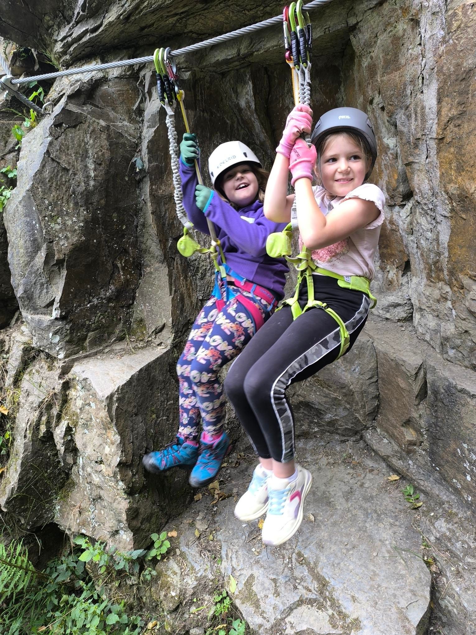 Zwei Kinder in Kletterausrüstung sind zu sehen, sie lachen. Sie hängen gesichert an ihrer Standplatzschlinge im Klettersteig.