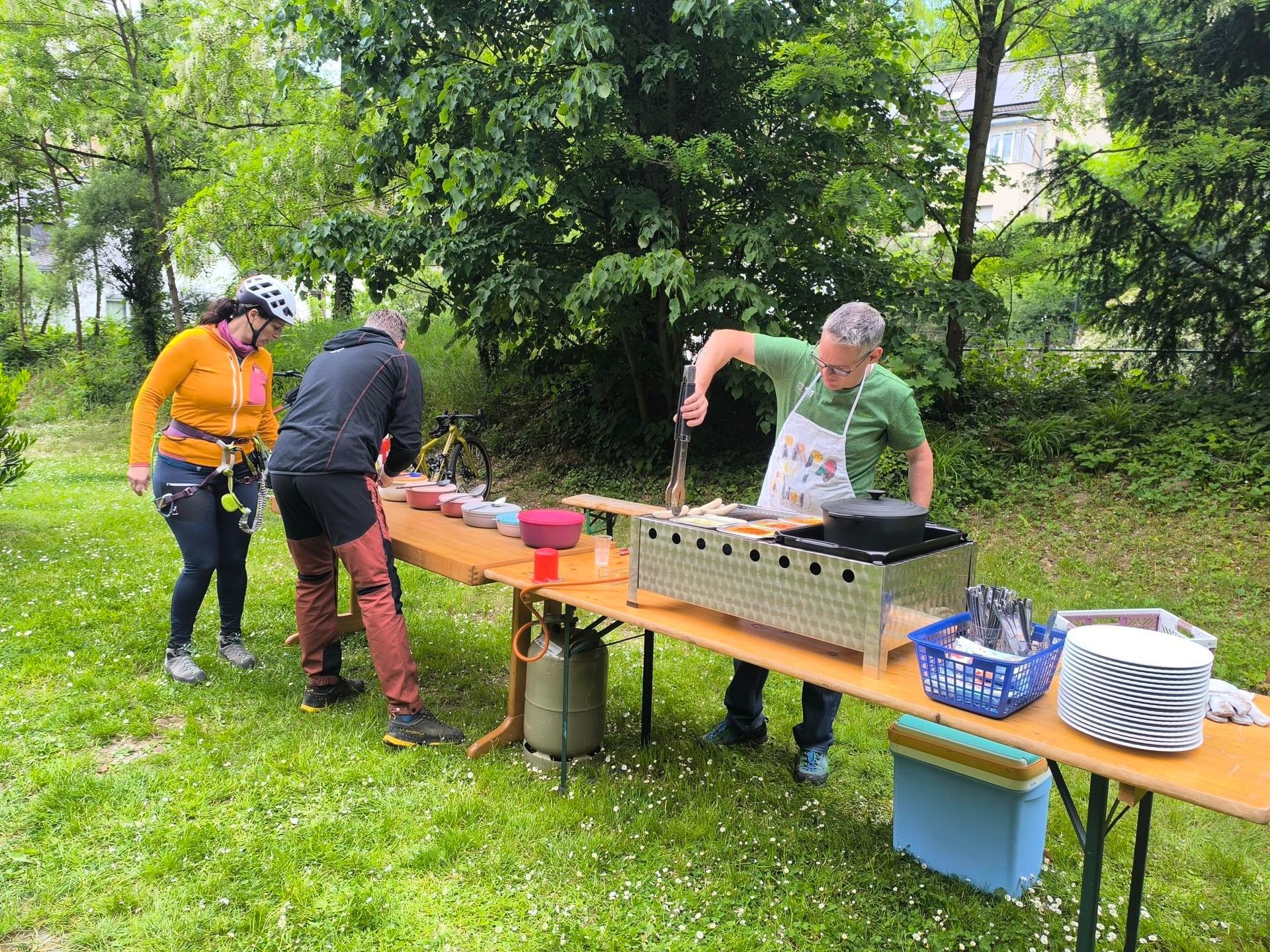 Eine Bierzeltgarnitur mit einem Grill ist zu sehen. Eine Person grillt, zwei weitere Personen bereiten auf einem weiteren Tisch Salate vor.
