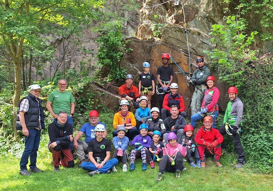 Ein Bild von einer Gruppe. 19 Personen tragen Kletterausrüstung mit Helm und Klettergurt. Links im Bild sind drei Personen, die keine Kletterausrüstung tragen.