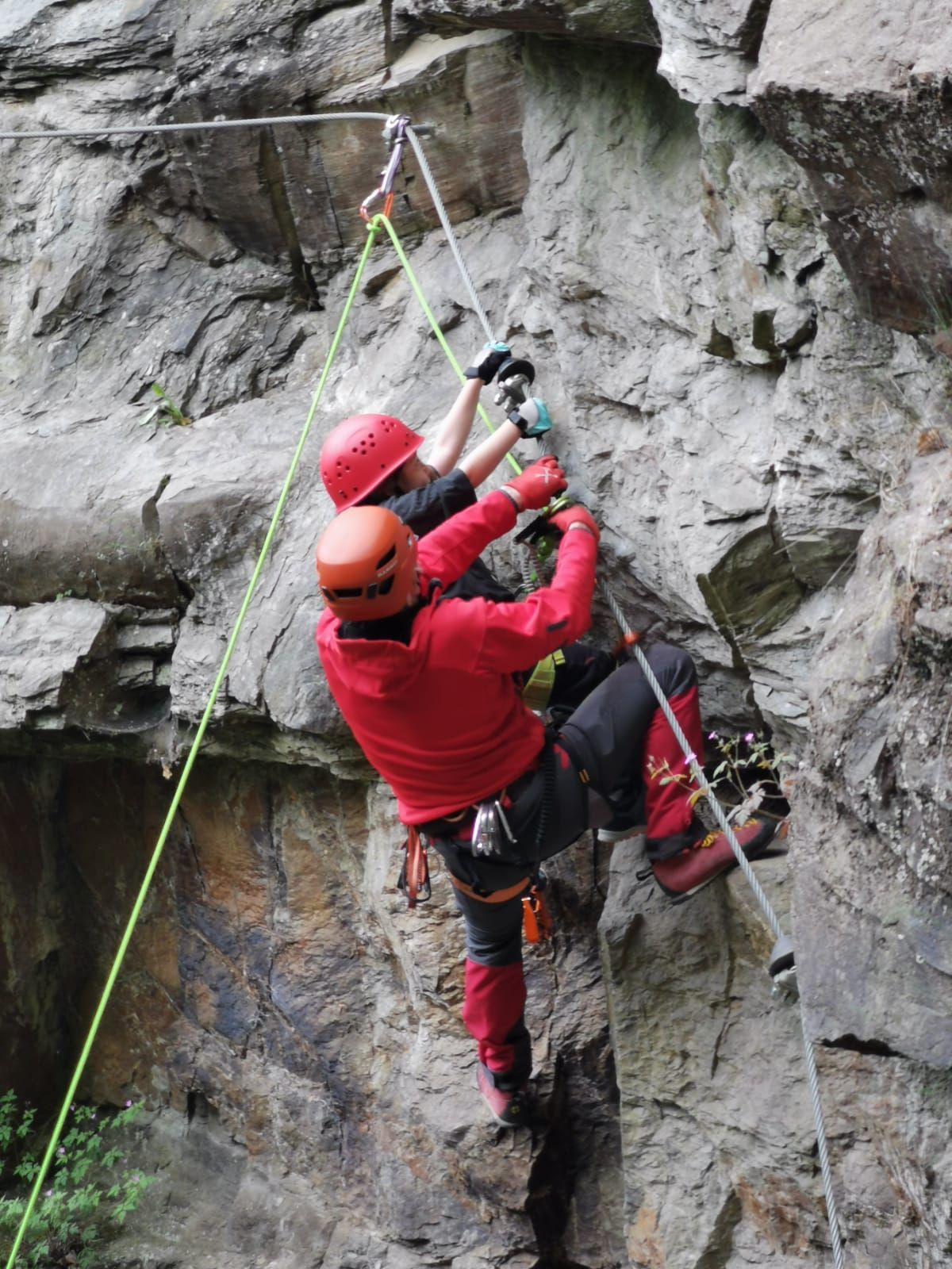 Zwei Menschen klettern an einem Klettersteig. Der hintere Person unterstützt die vordere Person beim Klettern.