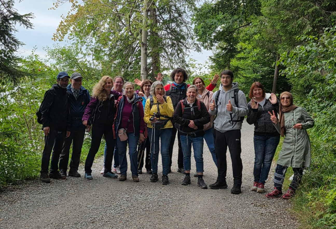 Gruppe von 14 Personen auf einem Kiesweg, umgeben von Bäumen und Sträuchern. Einige halten Wanderstöcke, andere posieren mit den Händen.