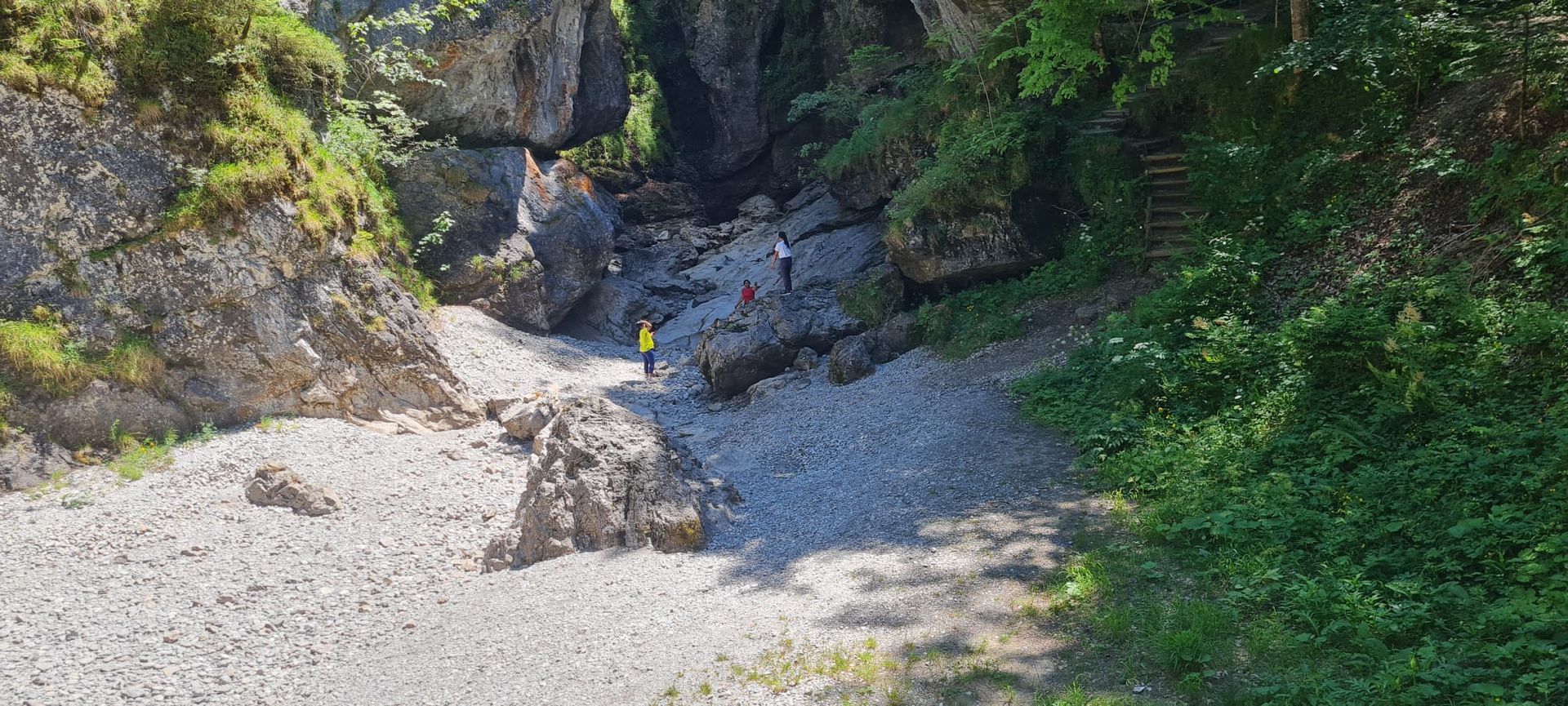 Schlucht mit großen Felsen und drei Personen auf einem kiesigen Weg, umgeben von Bäumen