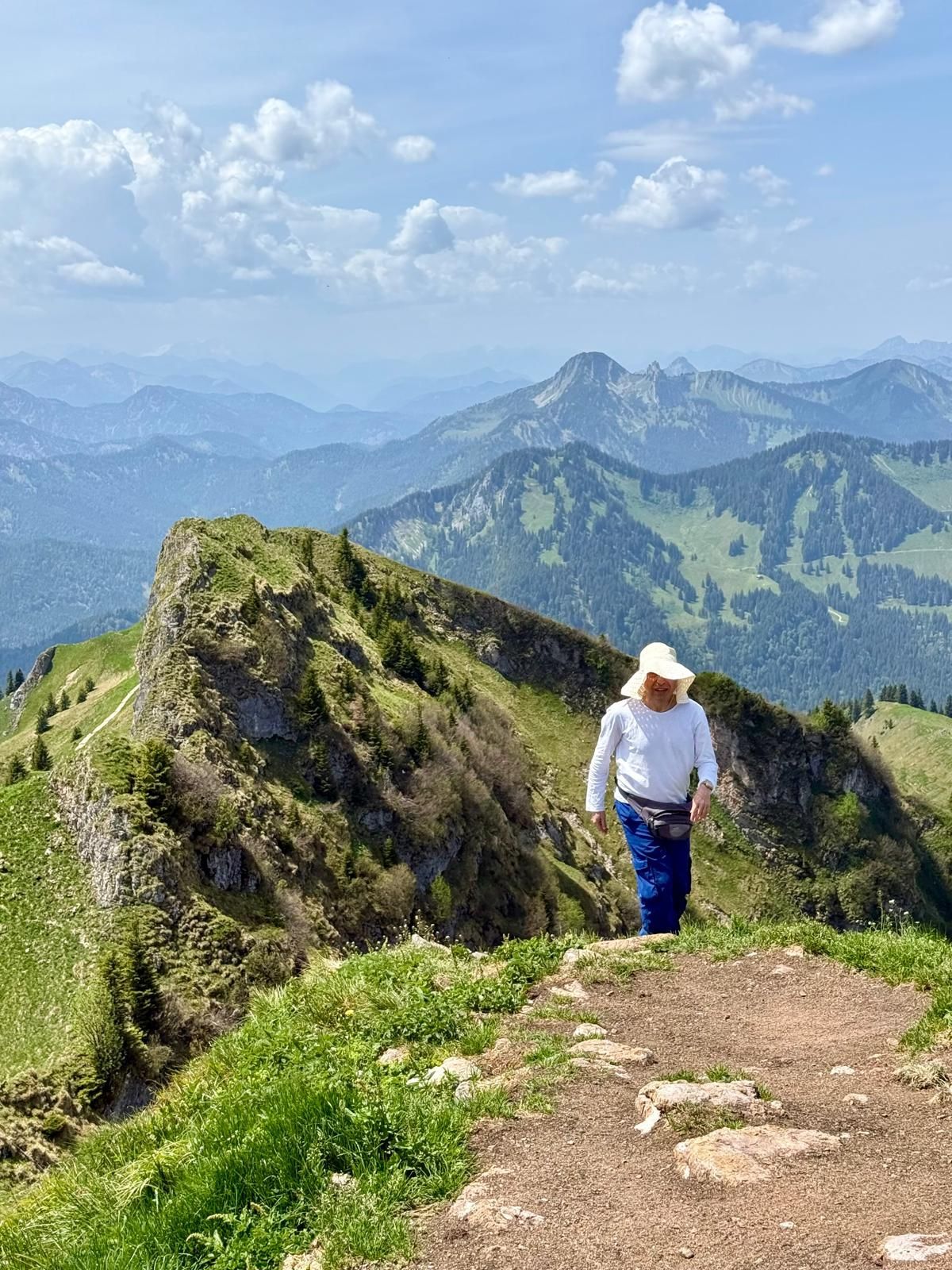 Wanderer mit weißem Hut und weißem Oberteil auf einem Bergpfad mit grünen Grasflächen und Bergpanorama im Hintergrund