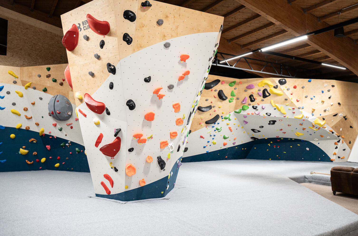 Boulderwand mit bunten Griffen in einer Kletterhalle, helle Holzwände und grauer Bodenbelag sichtbar.