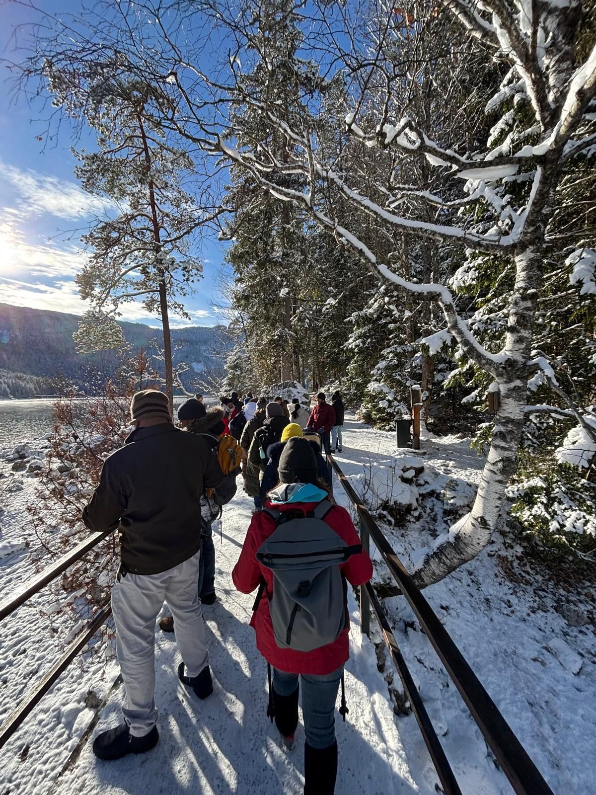 Eine Gruppe von Menschen in Winterkleidung wandert auf schneebedecktem Pfad entlang eines zugefrorenen Sees, umgeben von verschneiten Bäumen.