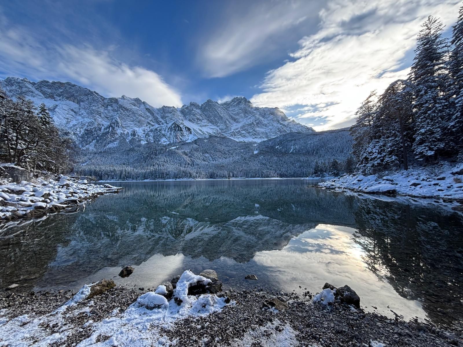 Eine schneebedeckte Bergkette spiegelt sich in klarem, ruhigem See unter blauem Himmel mit Wolken.