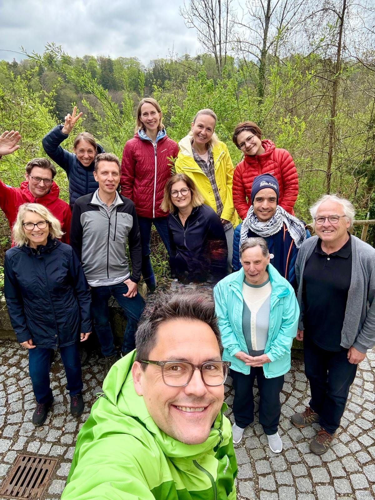 Gruppe von Personen in Outdoor-Kleidung steht auf gepflastertem Weg vor Bäumen und Sträuchern.