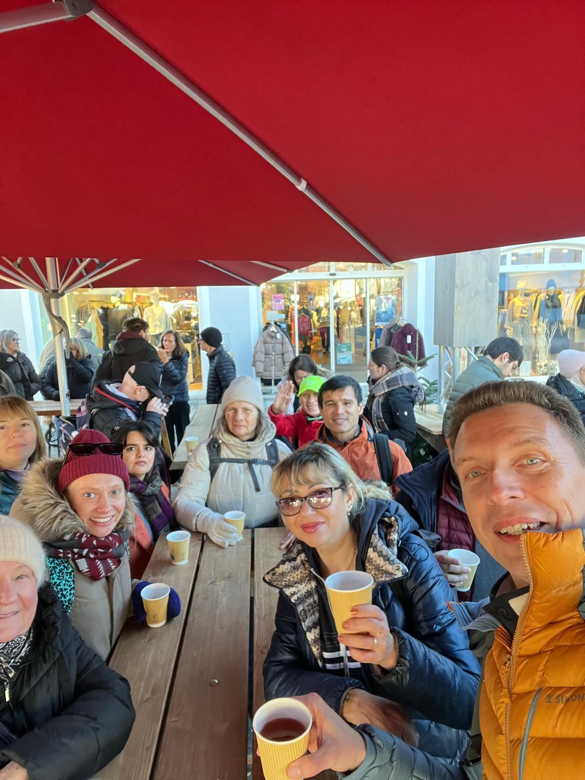 Gruppe von Menschen sitzt an einem langen Holztisch unter einem großen roten Sonnenschirm und hält Pappbecher in der Hand