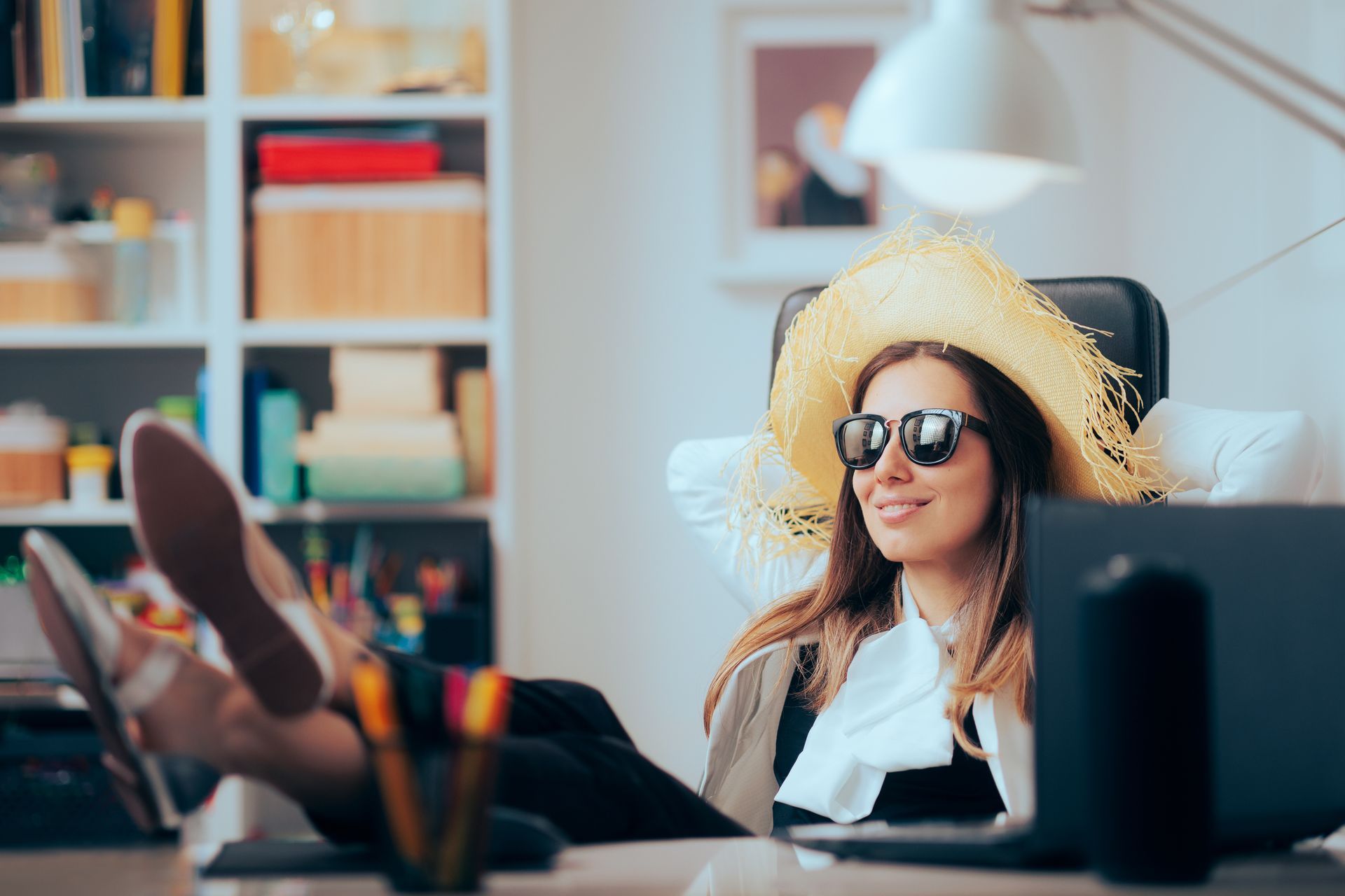 Eine Frau sitzt mit Sonnenbrille und Strohhut vor einem Bildschirm an einem Schreibtisch.
