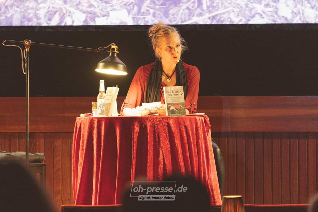 Nadine Witt liest aus ihrem Krimi Der Blaubart von Fehmarn vor einer großen Kino Leinwand. Bei abgedunkeltem Licht hörten die Interessierten Kinobesucher aufmerksam zu.