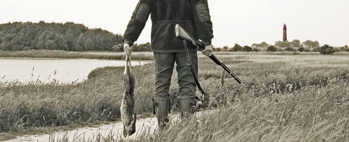 Ein Jäger auf dem Weg zum Flügger Leuchtturm Ein Jäger auf Fehmarn erlegt einen Hasen für die Buchgestaltung des Kriminalromans Der Blaubart von Fehmarn von Nadine Witt auf dem Deich mit Leuchtturm von Flügge im Hintergrund