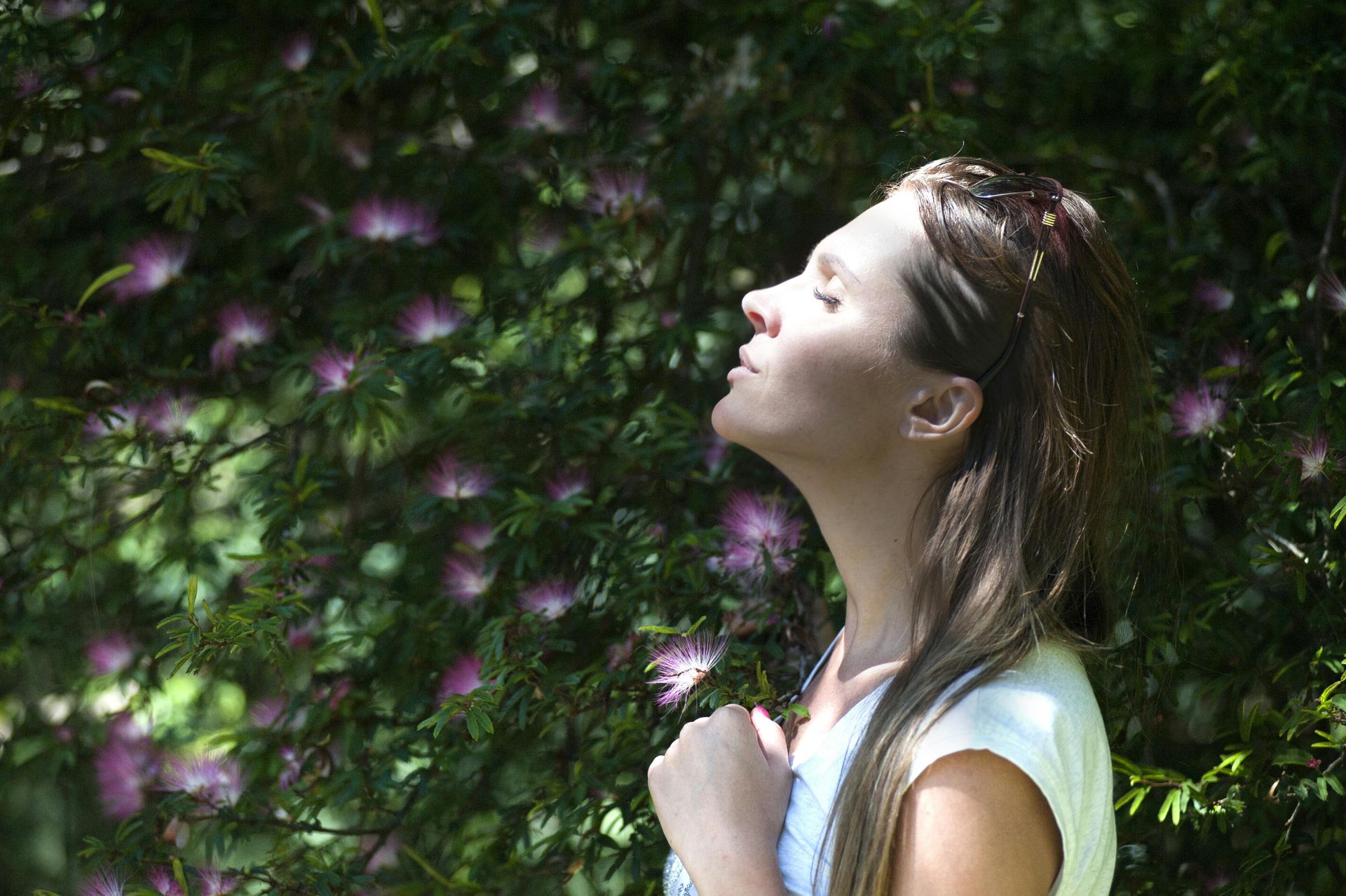 woman next to a hedge with purple flowers lifting head towards the sun with eyes closed