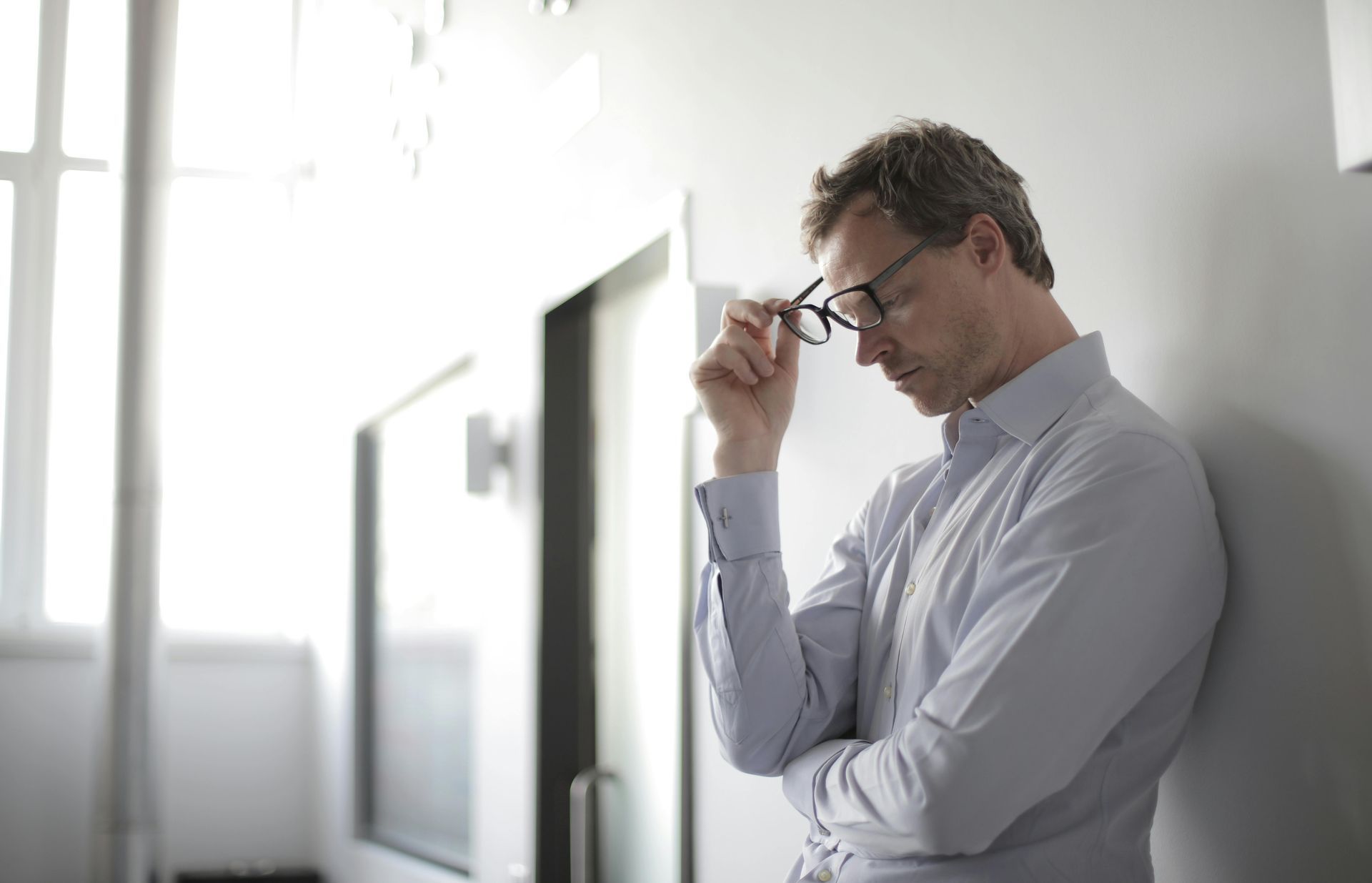Man in a shirt leaning against a wall taking glasses off