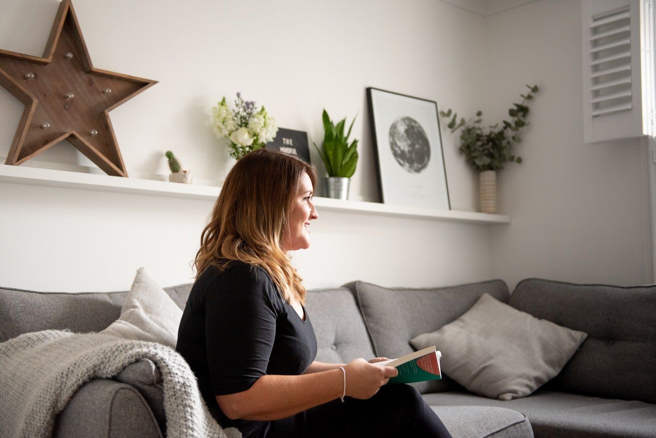 Photograph of a woman sitting on a sofa with a book open on her lap. Looking like she is in discussion with someone out of the shot.